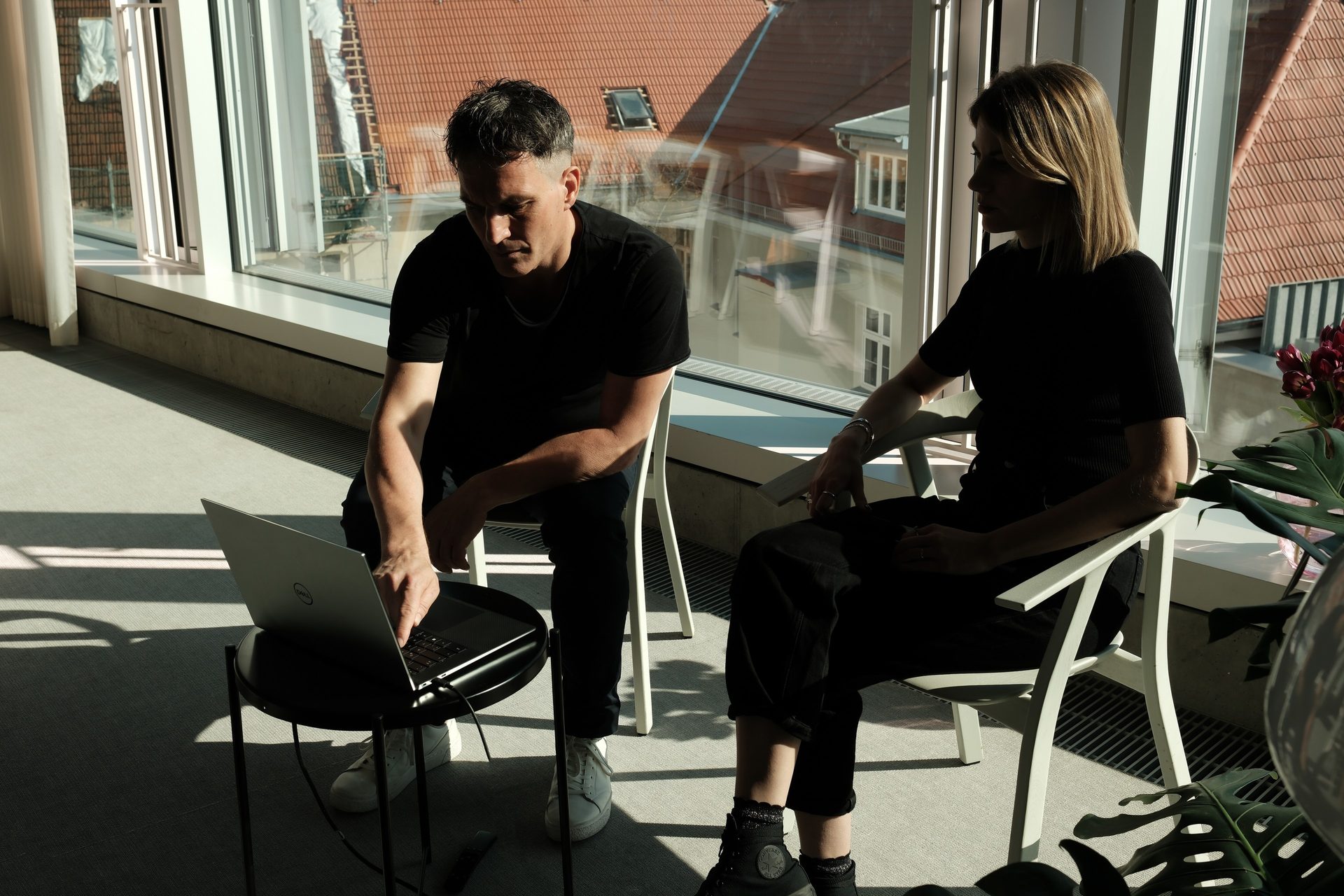 Zwei Personen im Gespräch an der Fensterfront, Laptop auf dem Schoß, Blick über die Dächer